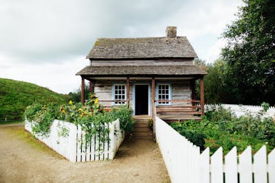 A quaint wooden long-term, rental house surrounded by greenery and a white picket fence,