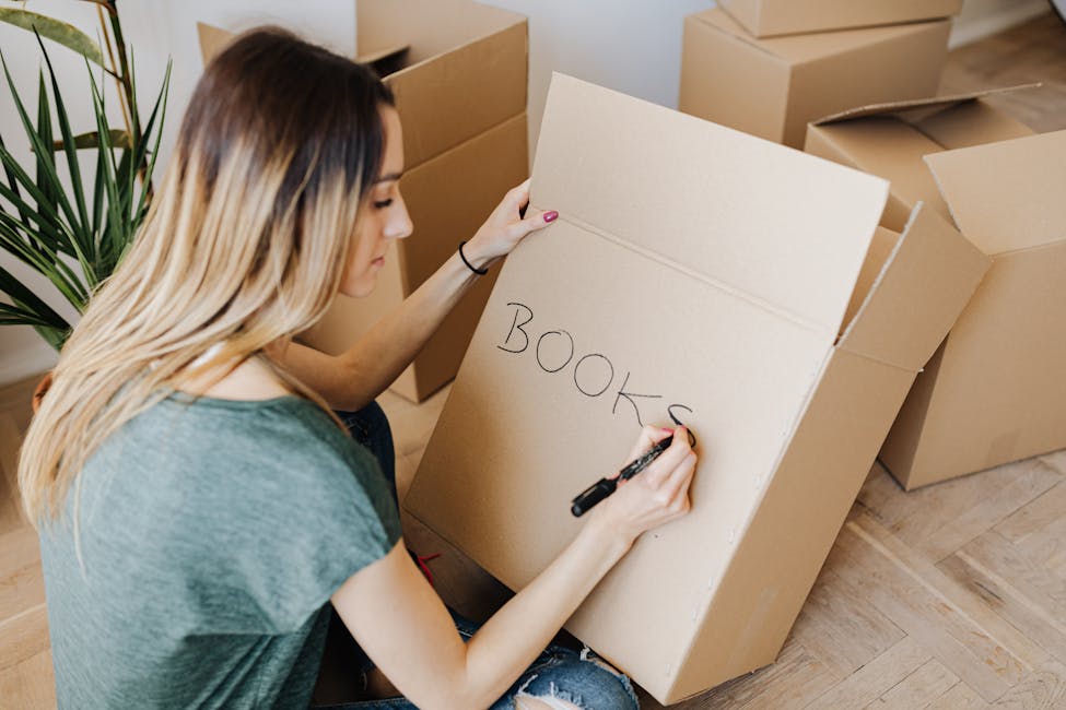 Young woman writing on cardboard box while packing books, preparing for moving as a tenant in a rental property.