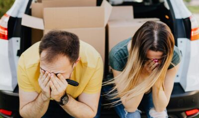 A distressed couple sits by a car full of boxes, showcasing the frustration behind hidden costs of corporate rental properties.