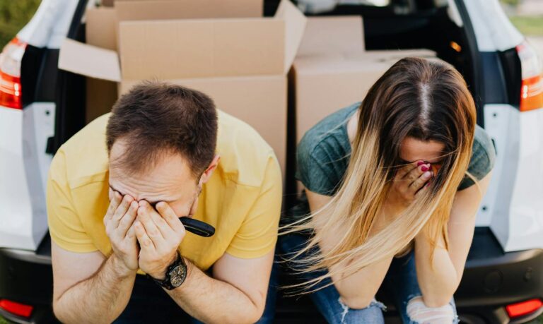 A distressed couple sits by a car full of boxes, showcasing the frustration behind hidden costs of corporate rental properties.