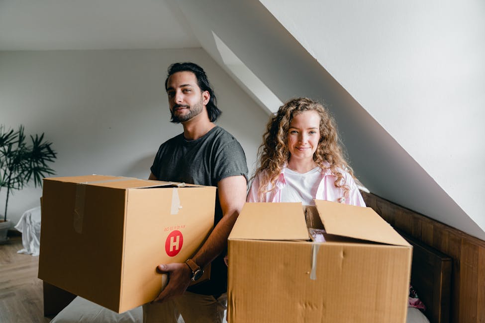Young woman and ethnic man in casual clothes smiling at camera and holding big boxes during relocation to new rental property showcasing the increase in rental costs.