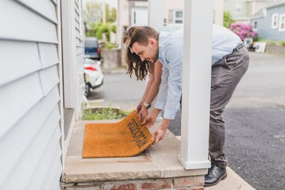 A young couple setting up a welcome mat on the porch of their new home, symbolizing how leverage, tax breaks, and long-term stability make property the ultimate wealth-building tool.