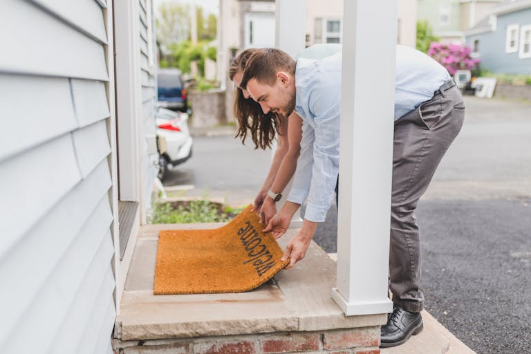 A young couple setting up a welcome mat on the porch of their new home, symbolizing how leverage, tax breaks, and long-term stability make property the ultimate wealth-building tool.