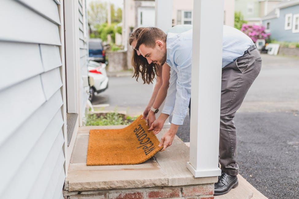 A young couple setting up a welcome mat on the porch of their new home, symbolizing how leverage, tax breaks, and long-term stability make property the ultimate wealth-building tool.