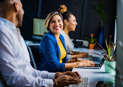 Three diverse professionals working and smiling at office desks, fostering teamwork and collaboration showcasing why Americans are no longer moving for work.