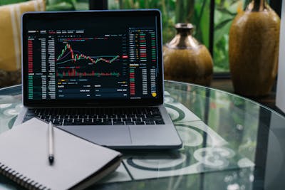 Laptop displaying cryptocurrency stocks and graphs on a glass table with a notepad showcasing real estate stocks.
